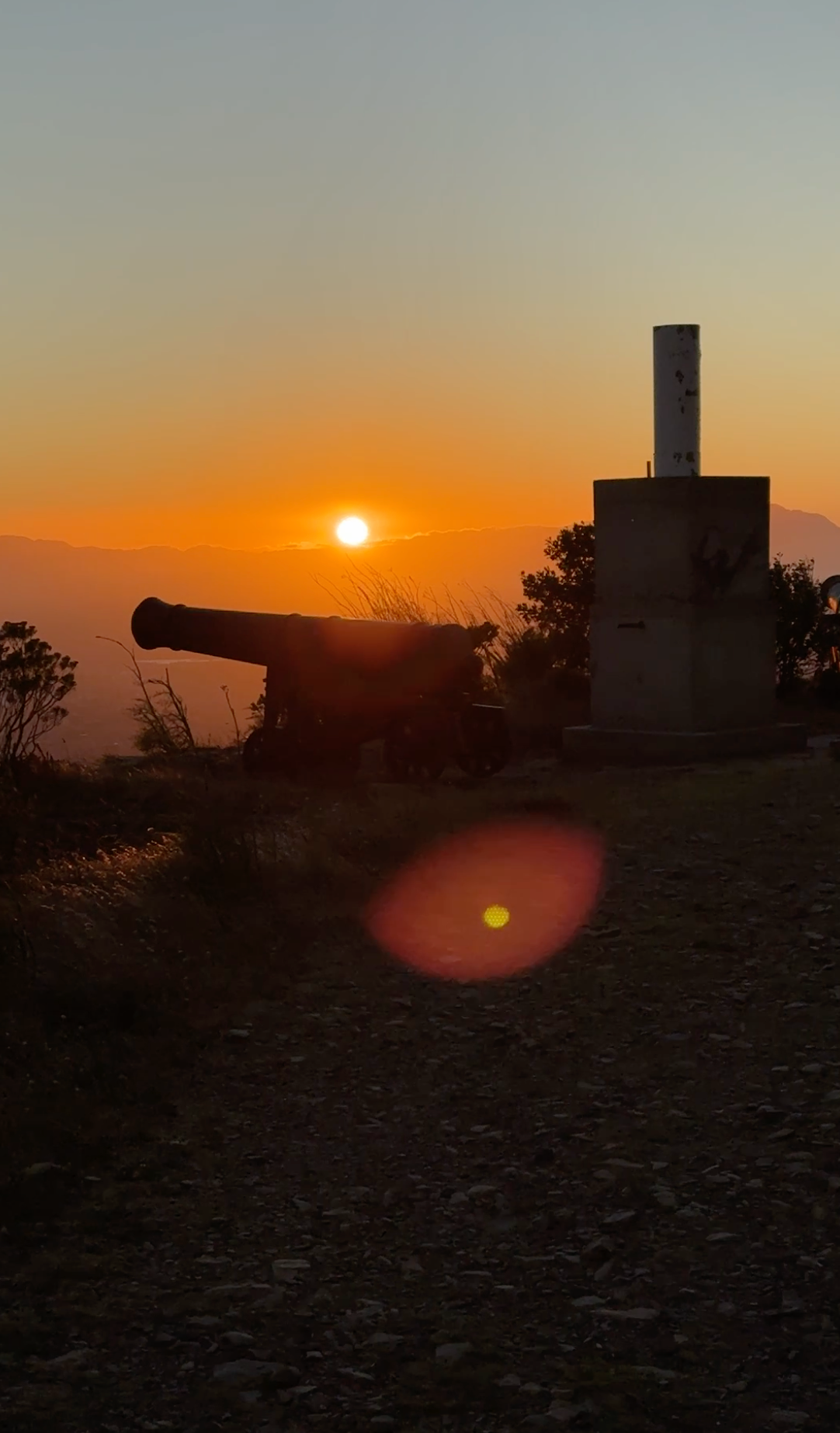 Sunset over a silhouetted cannon and tower, with orange sky and lens flare