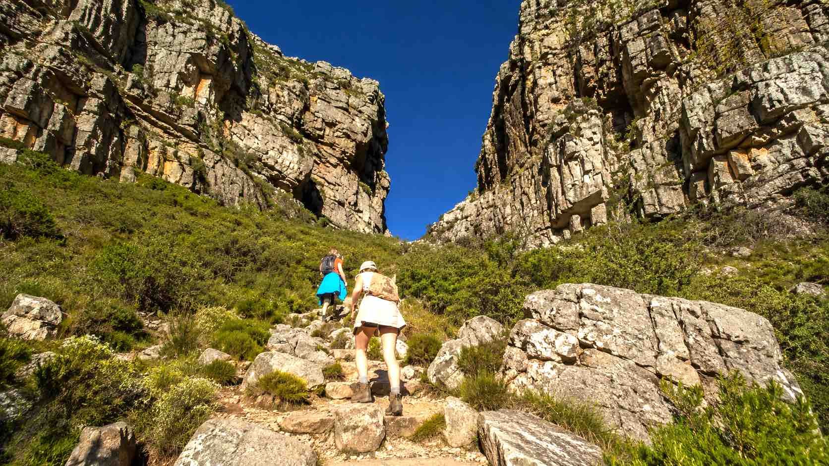 Two hikers ascending a rocky trail between steep cliffs under a clear blue sky