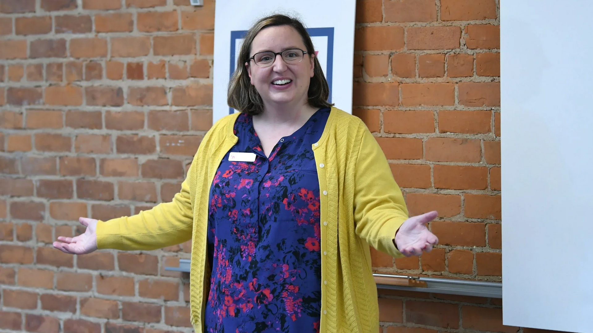 Woman with glasses and yellow cardigan, arms outstretched, smiling in front of a brick wall and sign.
