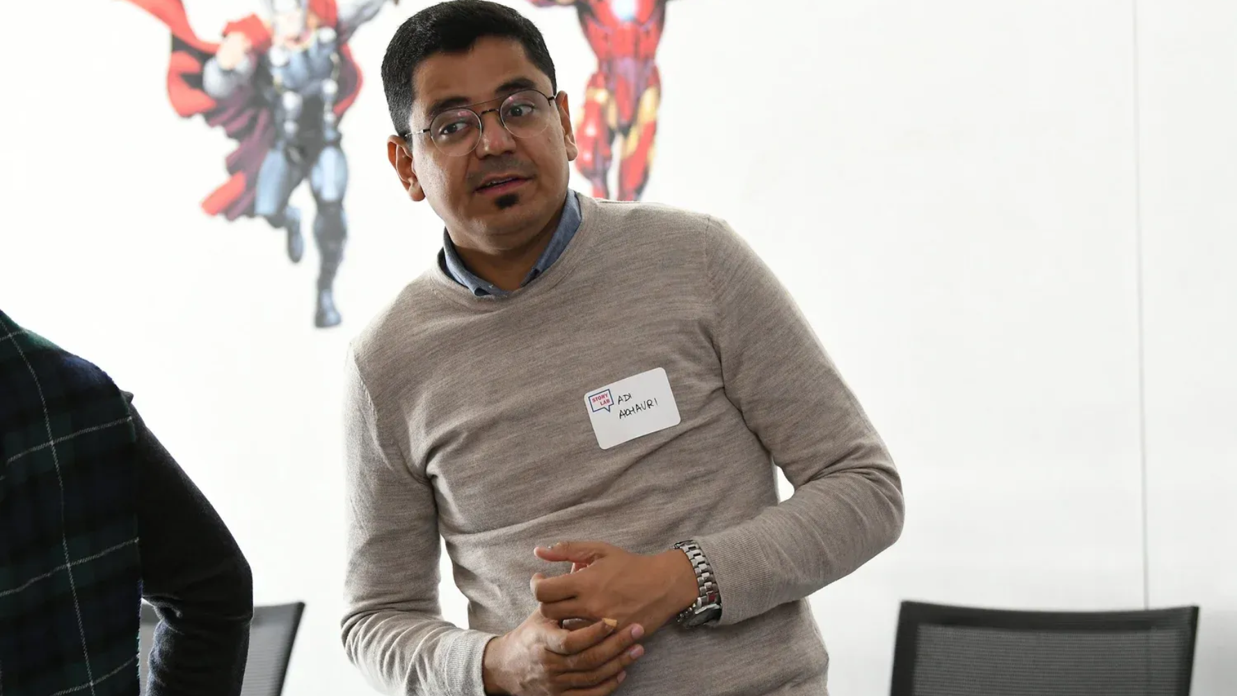 Man wearing glasses and sweater, gesturing in a meeting. Background with Marvel character art.
