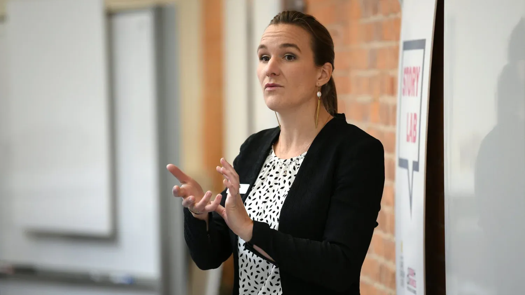 Woman gestures with hands while speaking in front of a white board and a brick wall.