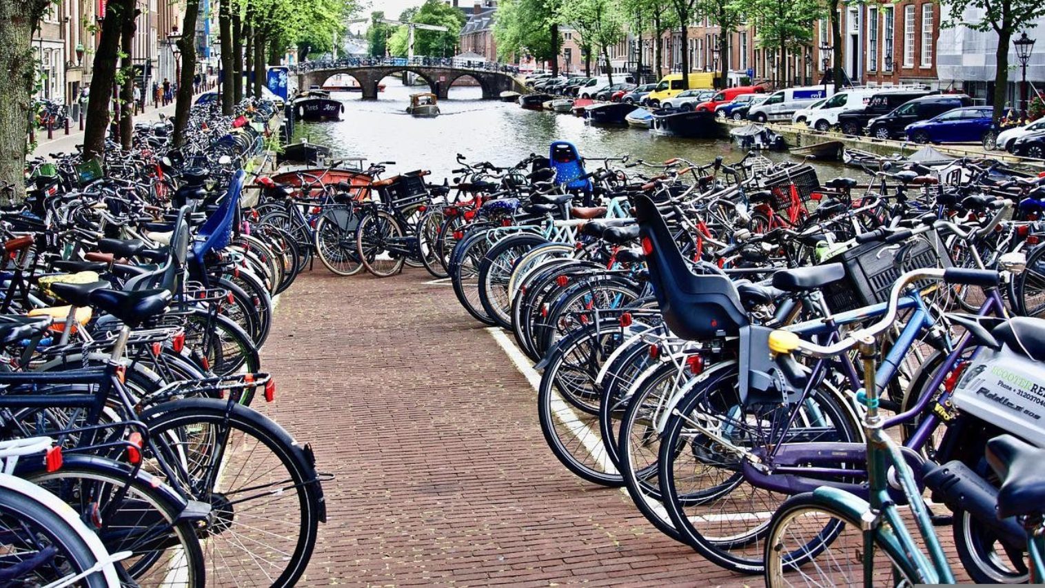 Rows of parked bicycles fill a brick-lined walkway next to a canal in Amsterdam; buildings and a bridge in the background.