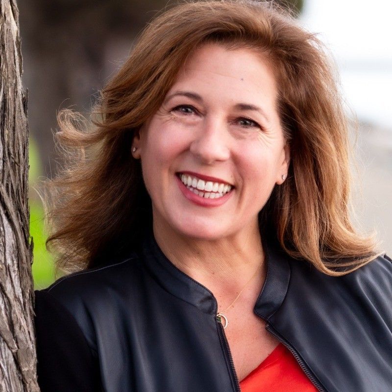 Woman smiles, leaning against a tree; wearing a black jacket and a red top.