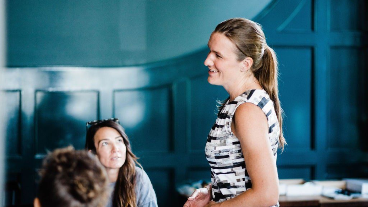 Woman speaking, smiling, in front of a group. Dark blue walls, natural light.