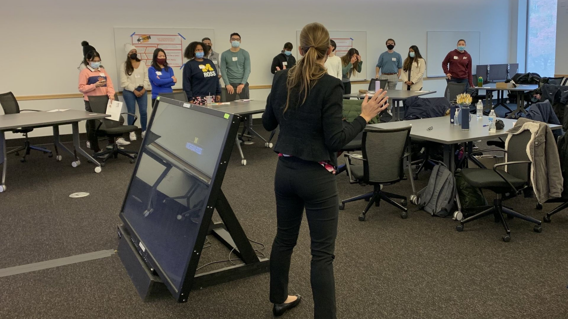 A woman speaks to a group of people standing in a classroom; a large screen sits in front of her.