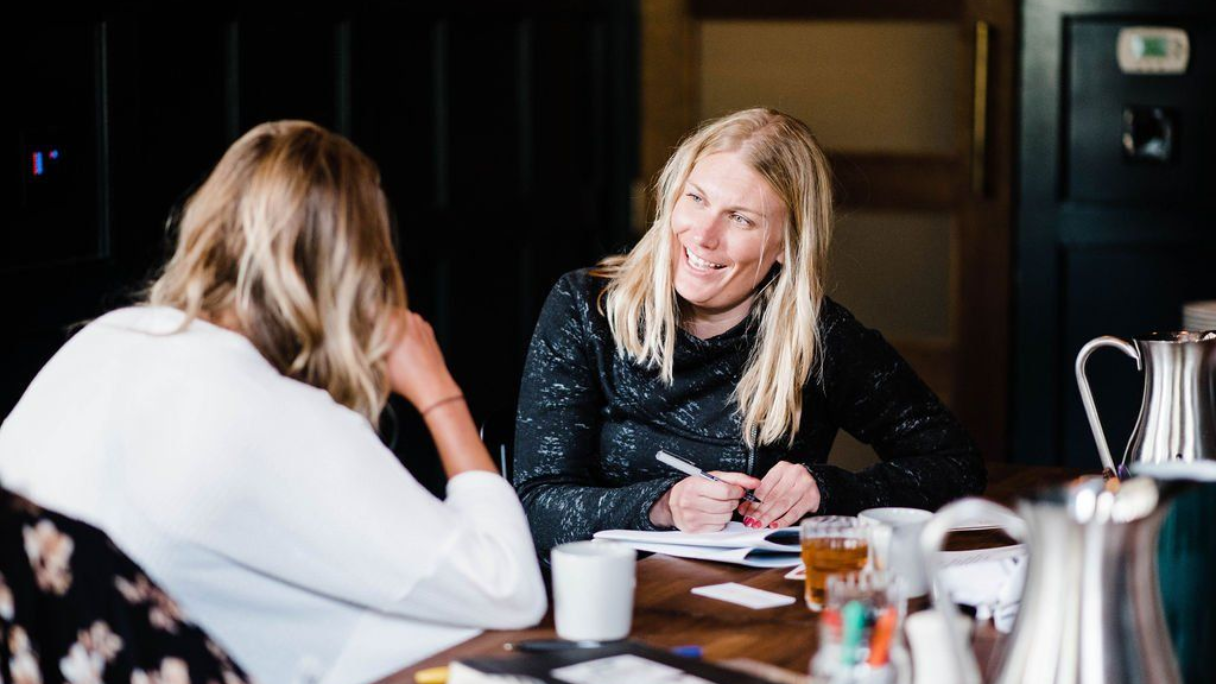 Two women at a table, one smiling and taking notes. A teapot, cup, and pens sit on the table.