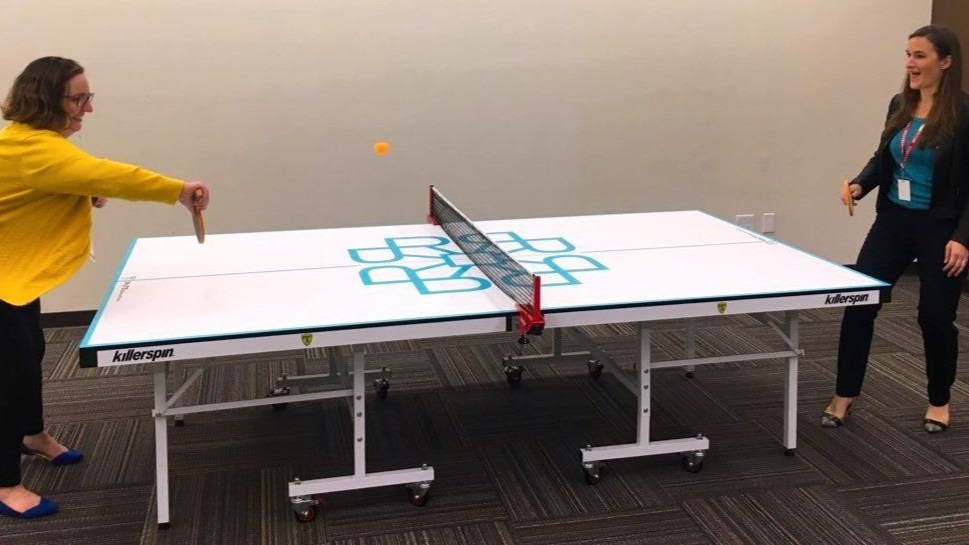 Two people playing ping pong in a room. One person swings a paddle, the other smiles, both stand near the white table with a blue design.