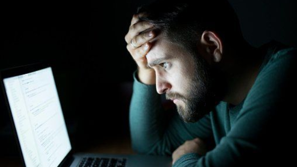 Man looking stressed at a laptop screen in the dark, hand on forehead.