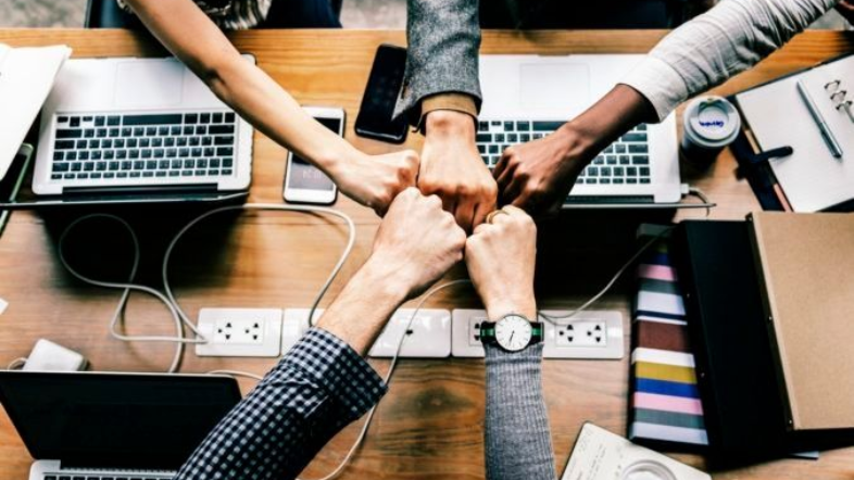 Hands bumping fists over a desk with laptops and office supplies.