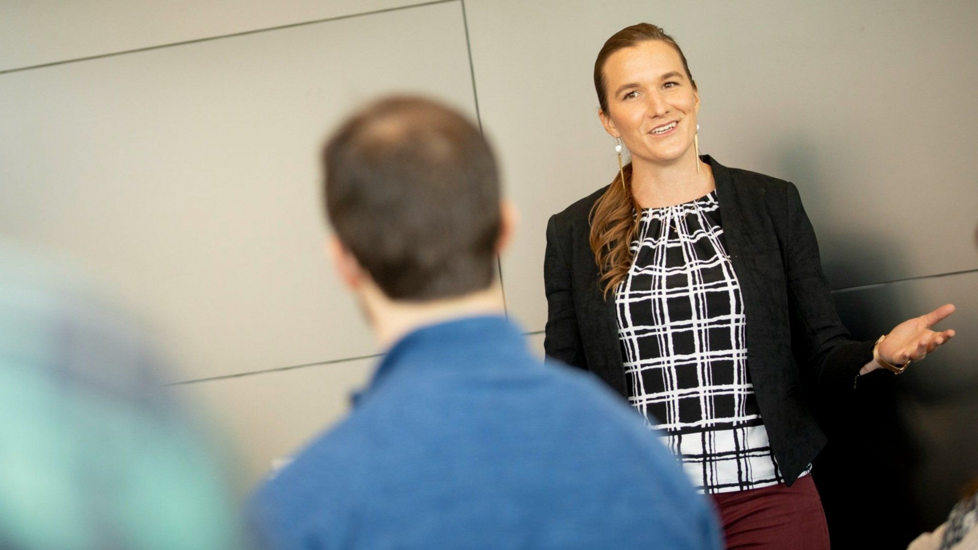 Woman speaking to a group, smiling, in front of a gray wall; wearing blazer, patterned top, and burgundy pants.