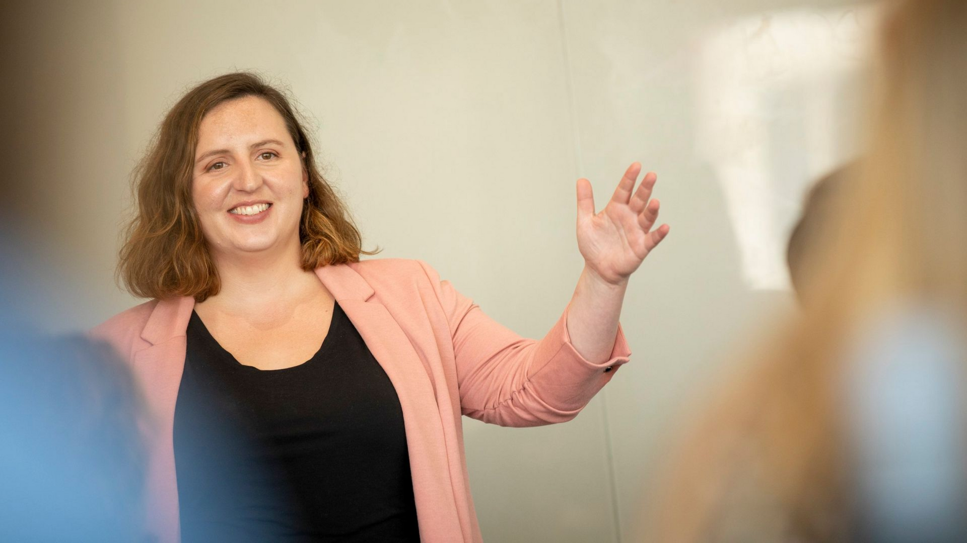 Woman in a pink blazer speaks and gestures to her right in a meeting.
