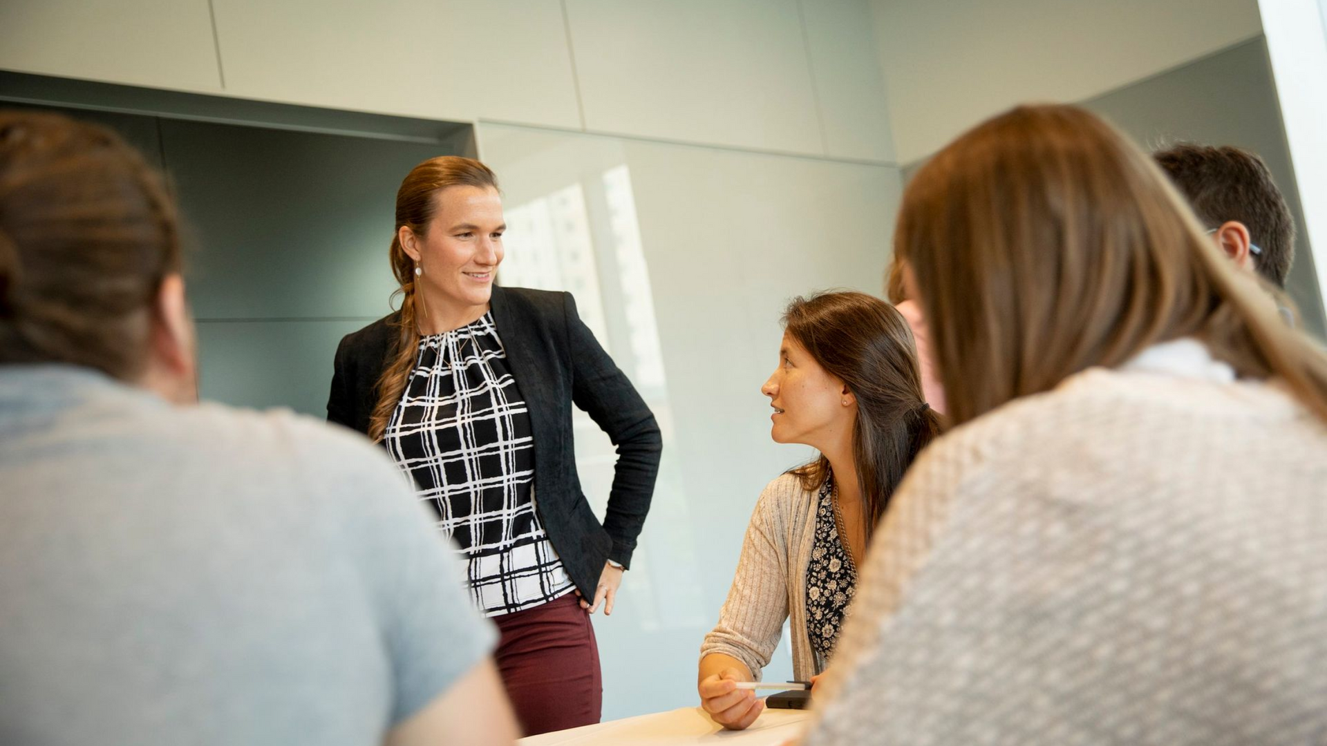 Woman in blazer leading a meeting, smiling. Others seated at a table. Bright room.