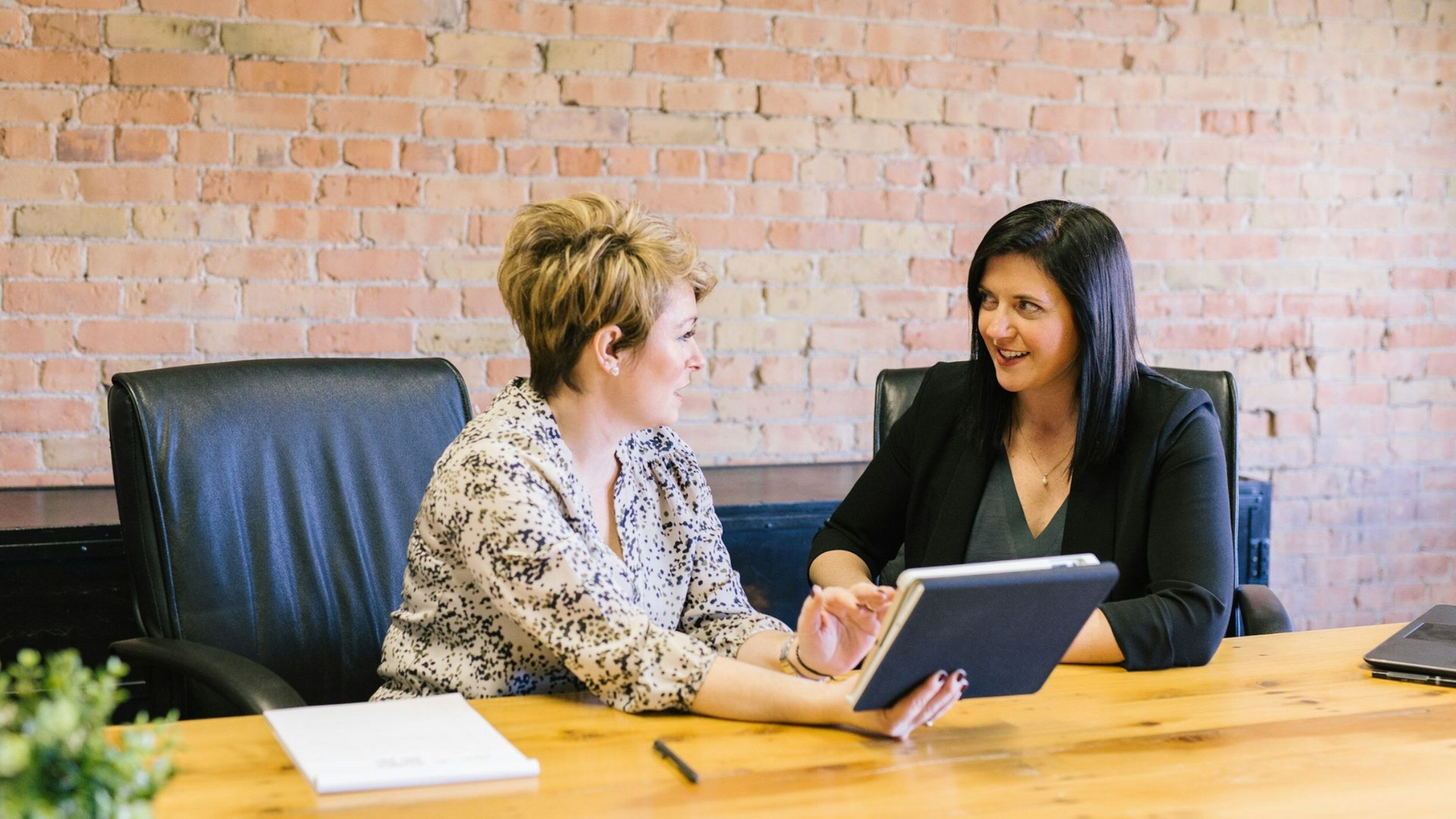 Two women at a wooden table, looking at a tablet, in an office with a brick wall.