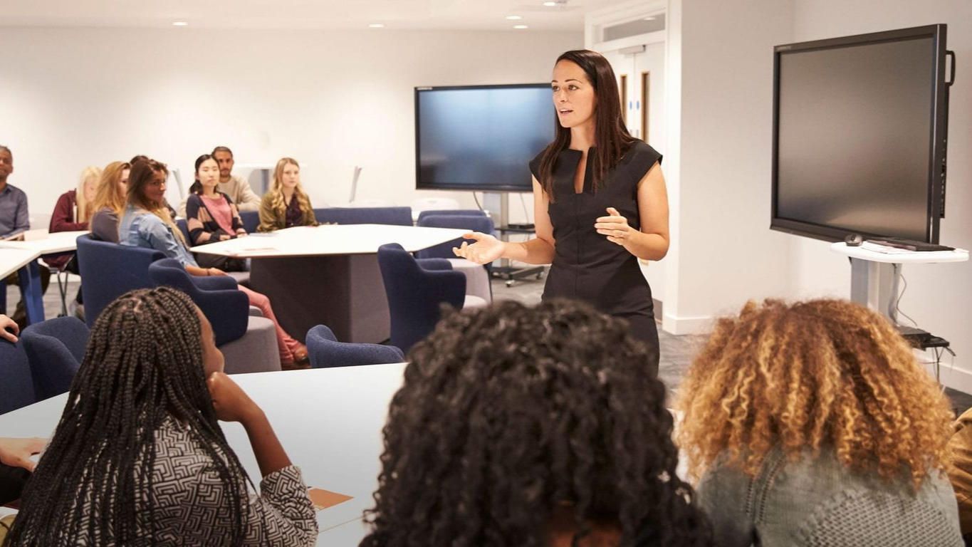 Woman presenting in a modern conference room; audience seated at a table.