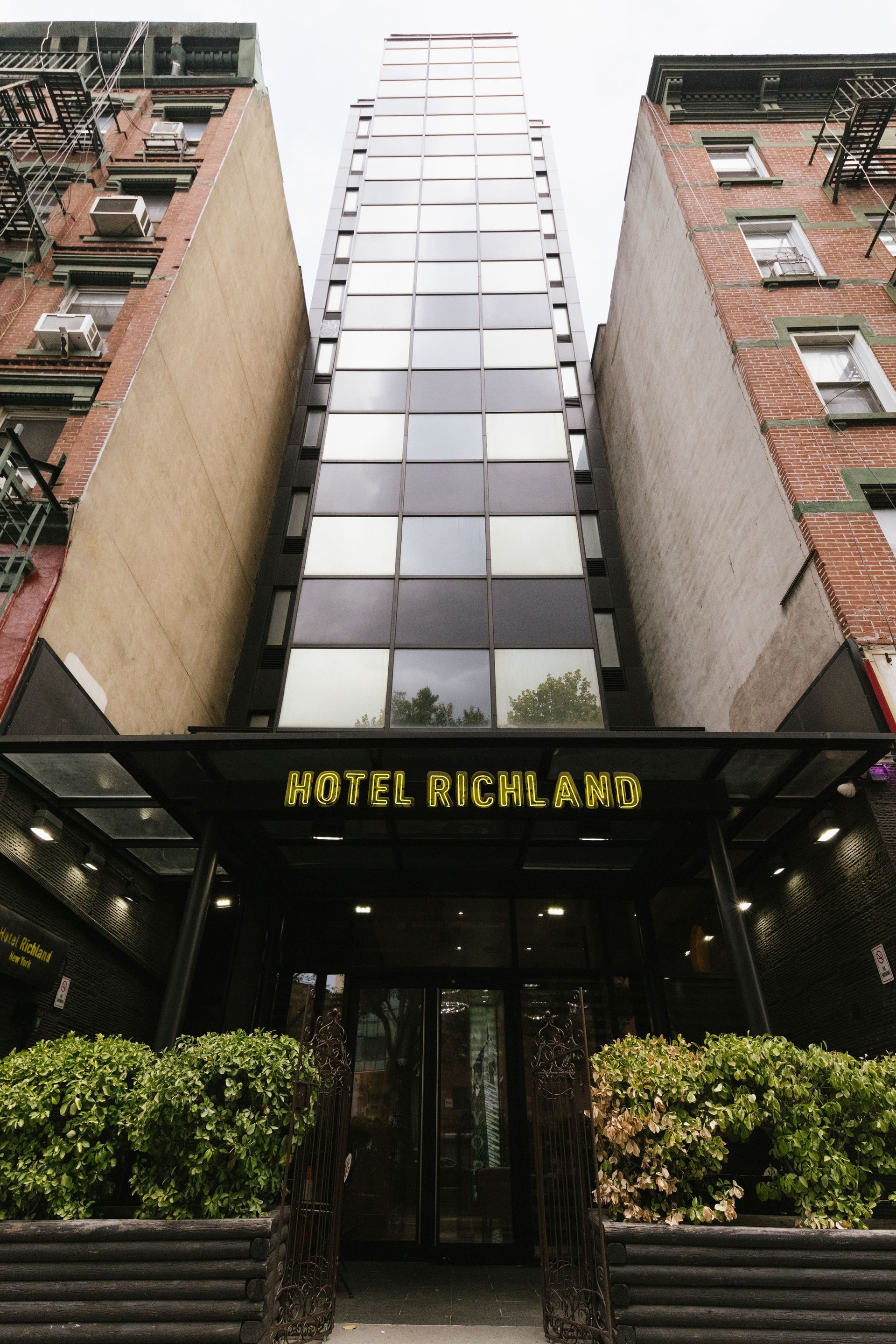 Hotel Highland entrance framed by buildings, glass facade rising high, with sign and planters.