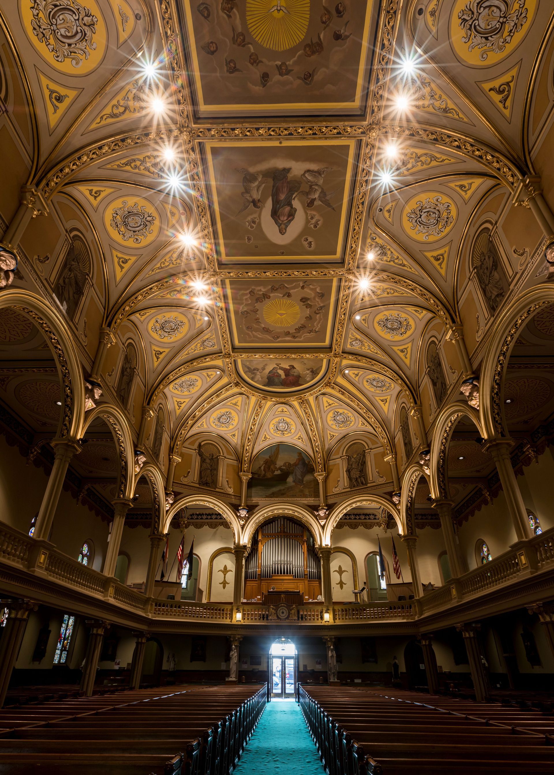 Ornate gold-and-cream cathedral interior with vaulted ceiling, chandeliers, and rows of pews leading to a bright doorway