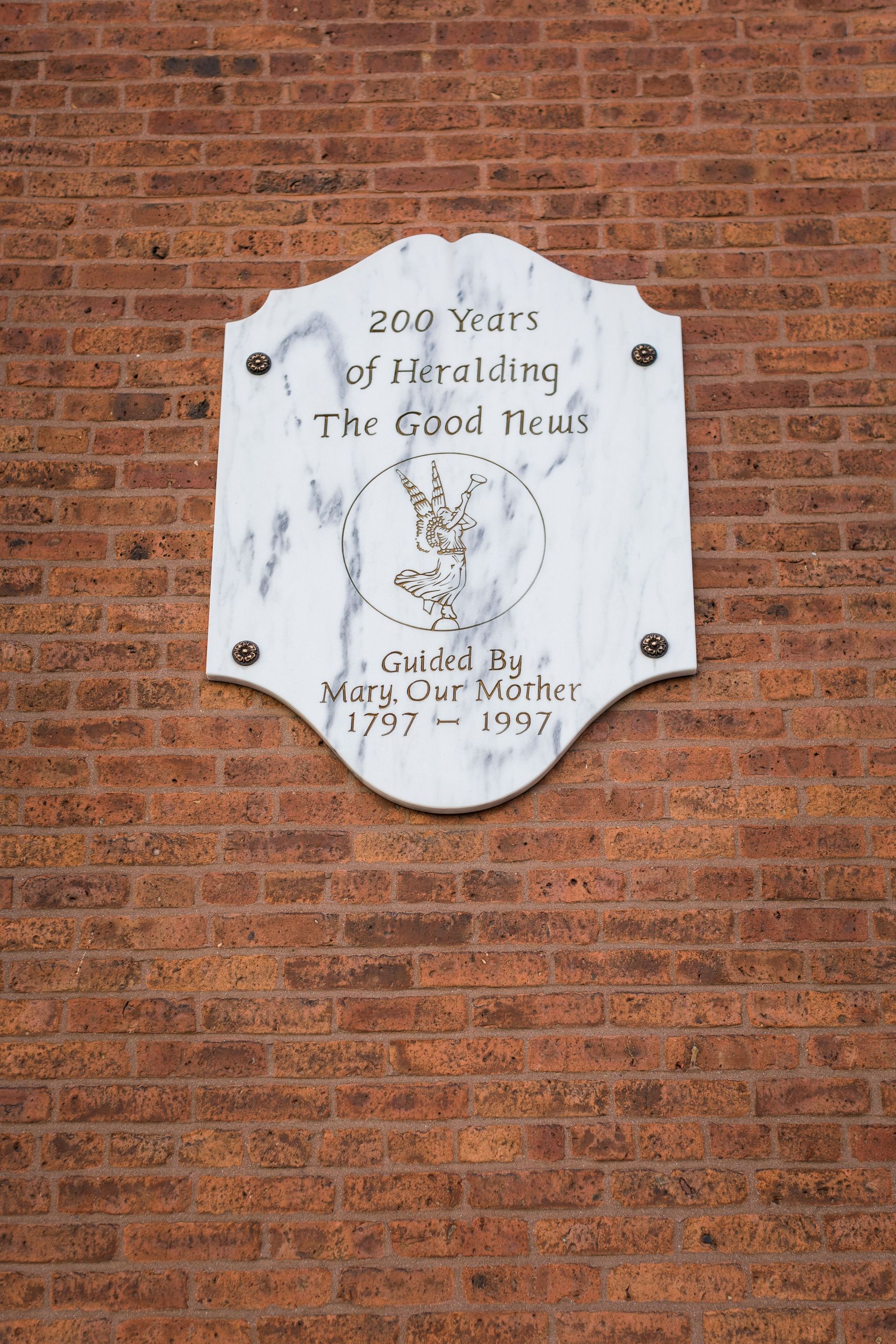 White commemorative plaque on a red brick wall with faded text and a gold seal at center.