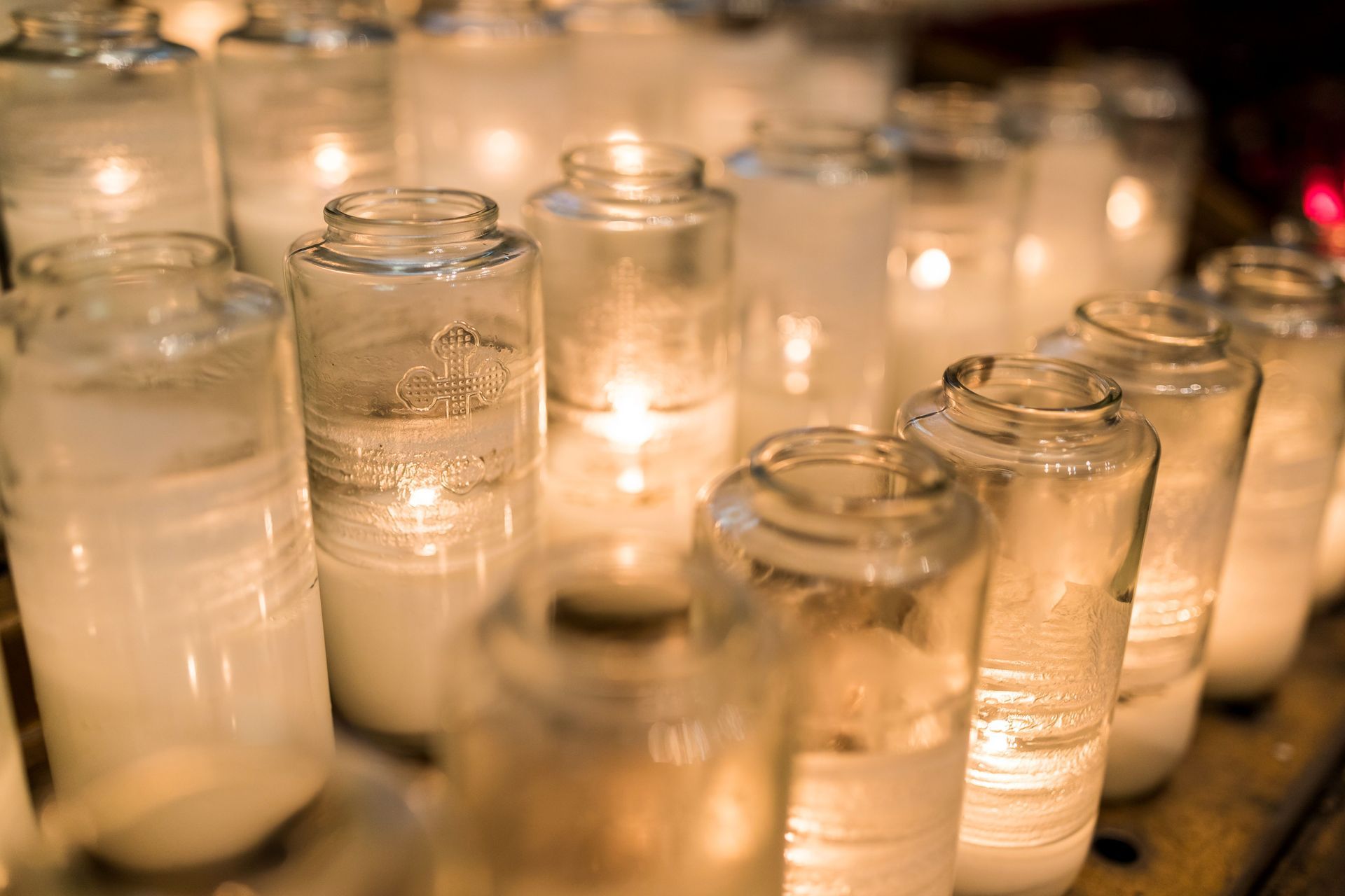 Rows of lit white candles in glass jars, softly glowing in a dim setting