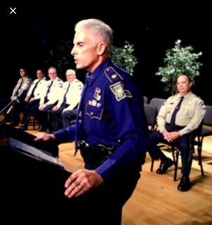 Uniformed officer speaking at a podium with seated officers behind him on a stage