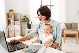 Woman working on laptop while holding a baby, both smiling. They are in a well-lit living room.