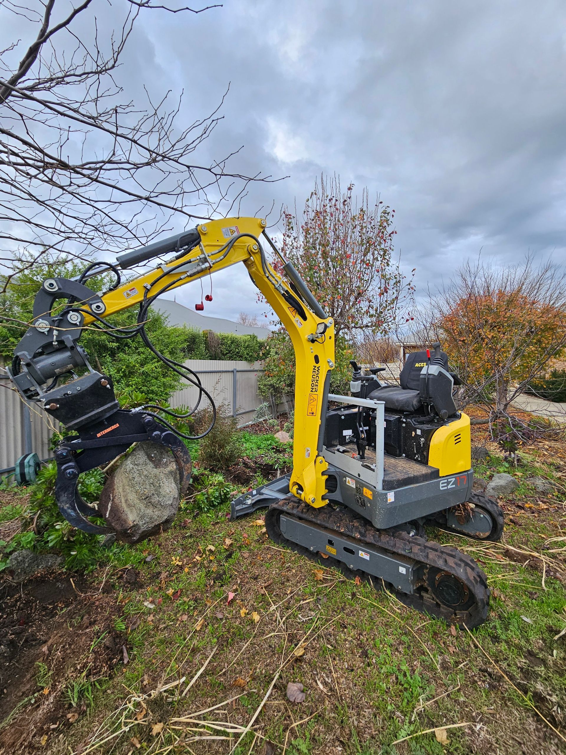 A small yellow excavator is cutting a tree stump in a yard.