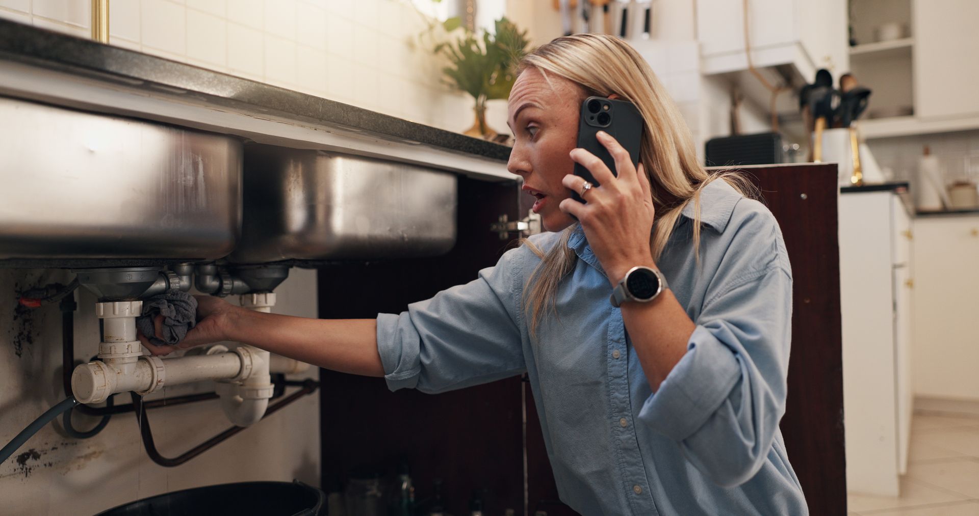 View of a blond woman kneeling next to a sink using a cellphone to call a plumber. View of a blond woman kneeling next to a sink using a cellphone to call a plumber.