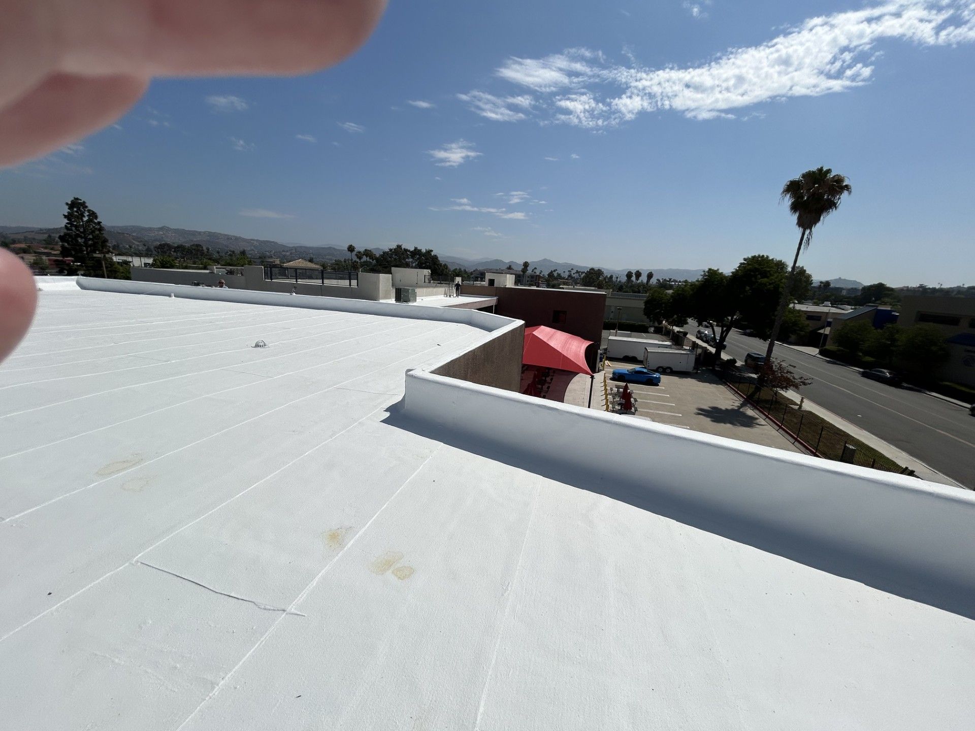 White rooftop with a red vent under a blue sky, viewed over a street with palm trees