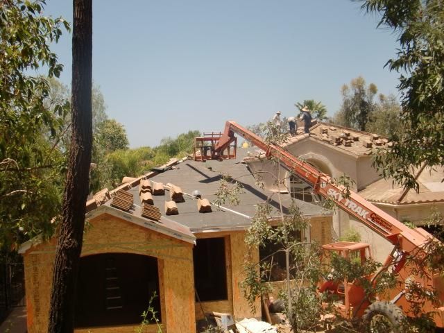 Roofers working on a house roof with a red crane lifting materials nearby, surrounded by trees.