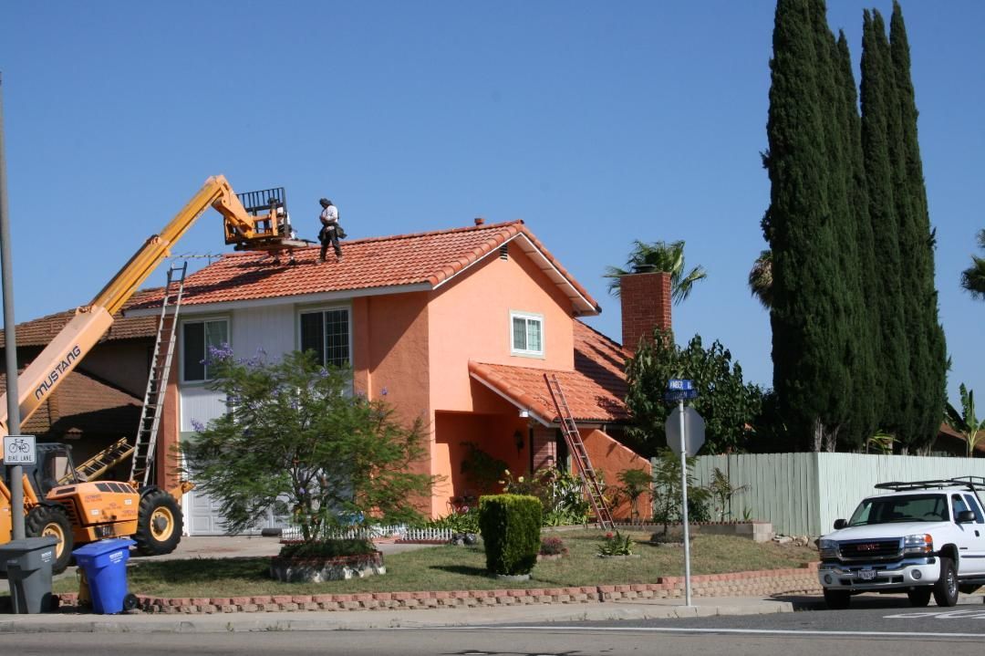 Workers on a yellow lift repairing a red-roofed house beside a street, with a white van nearby.