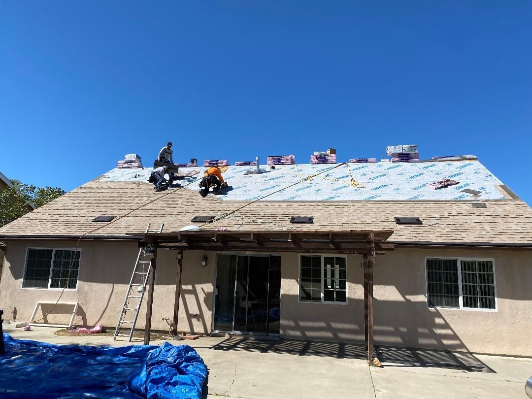 Workers on a roof repairing a house with blue tarp and ladders under a clear sky
