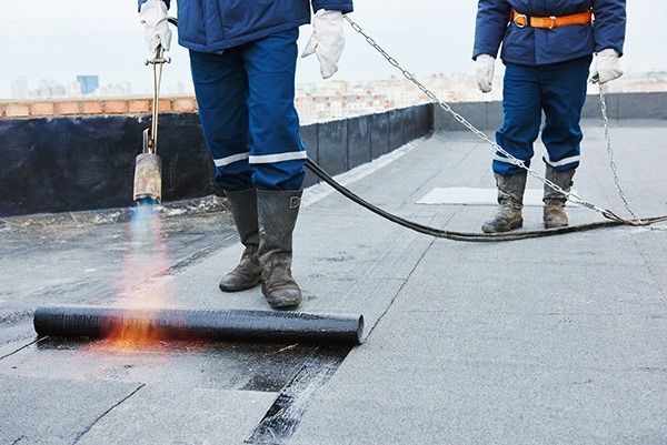 Workers torching a roof membrane during roofing repair, with visible flame and tools.