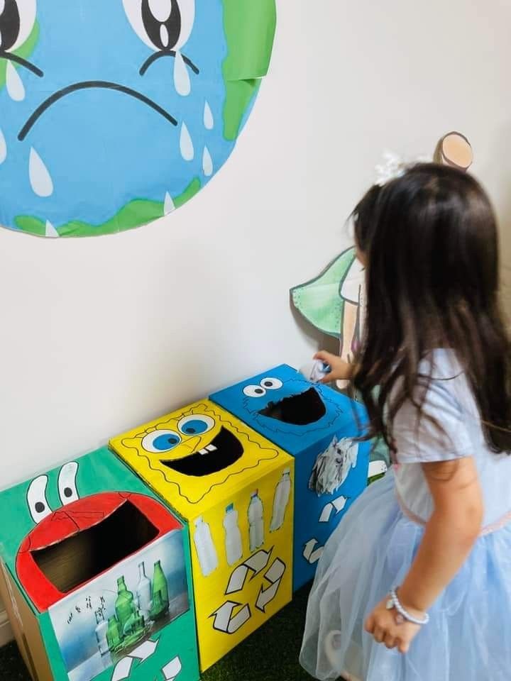 A child placing trash into one of three character-themed recycling bins, with a sad Earth drawing on the wall.