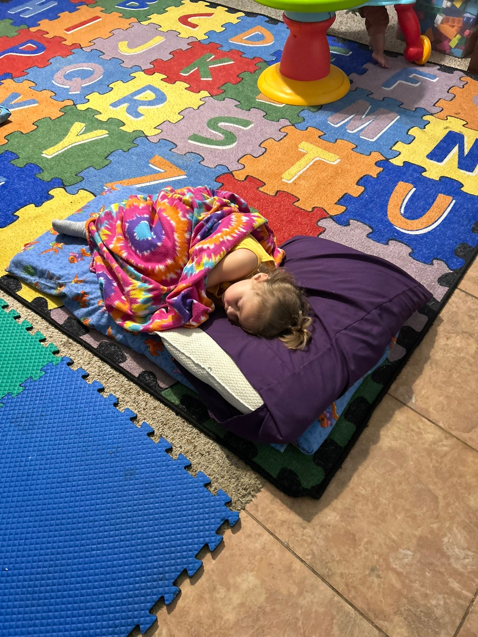 Childcare room with toys, table, and mats on tile and wood flooring. Brown walls and a white door.