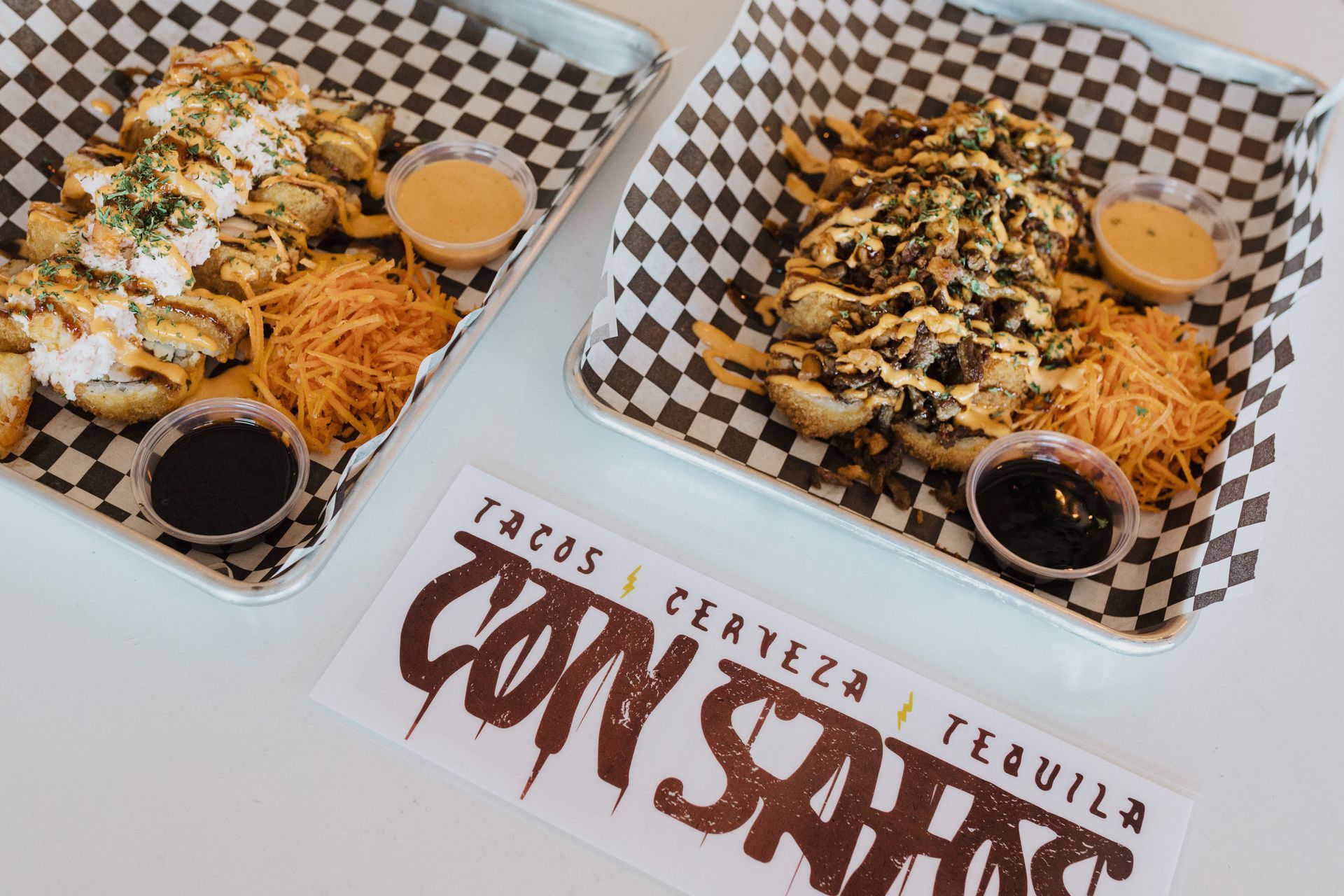 Two trays of food are sitting on a checkered paper on a table at con safos in denver colorado