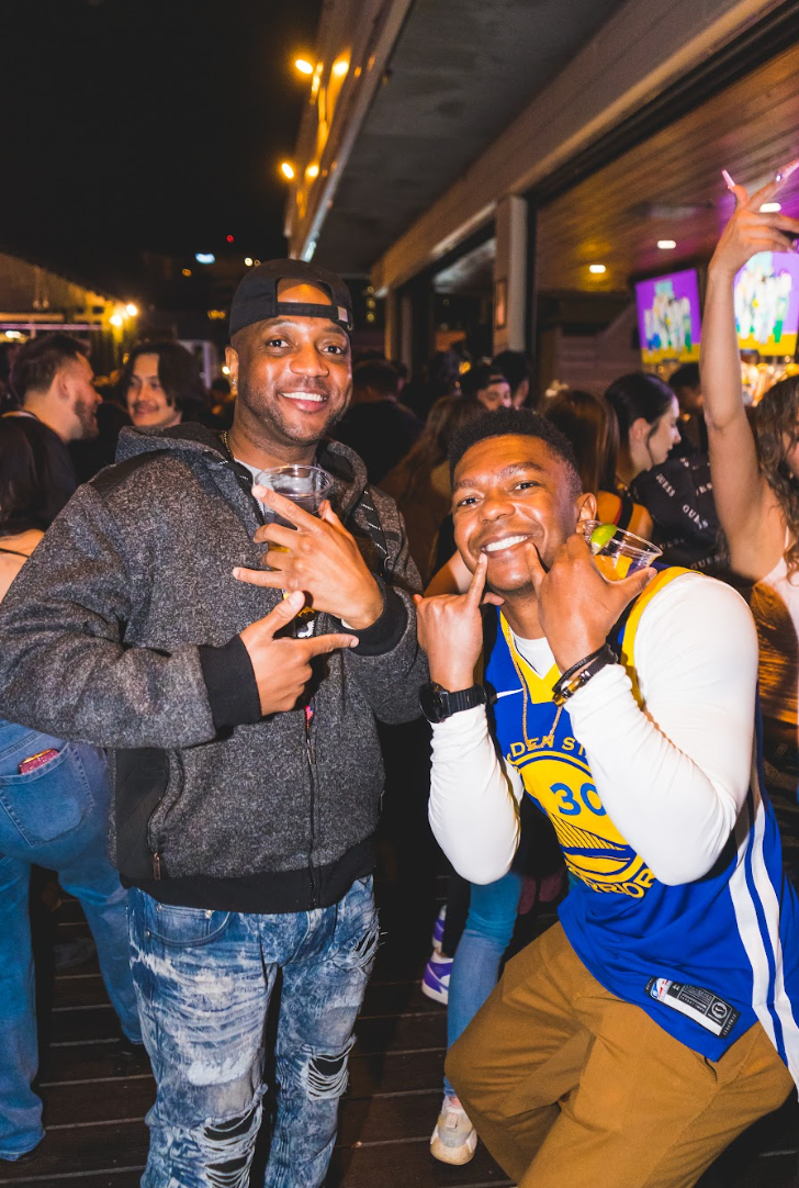Two men are posing for a picture at a party . one of the men is wearing a golden state warriors jersey. they are located at el patio in denver colorado