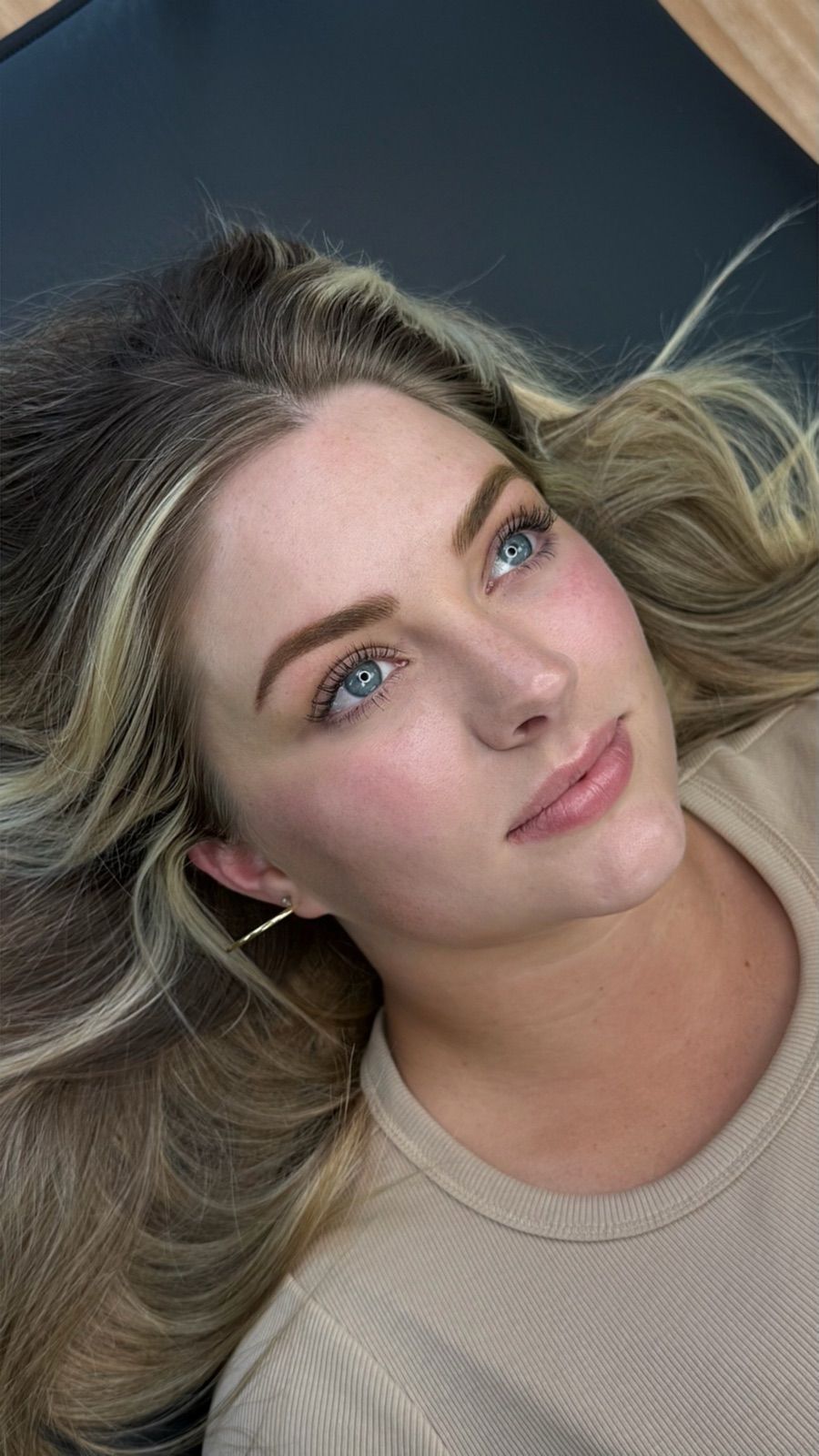 Close-up of a woman with wavy blonde hair and blue eyes, wearing a beige top, lying on a dark surface.
