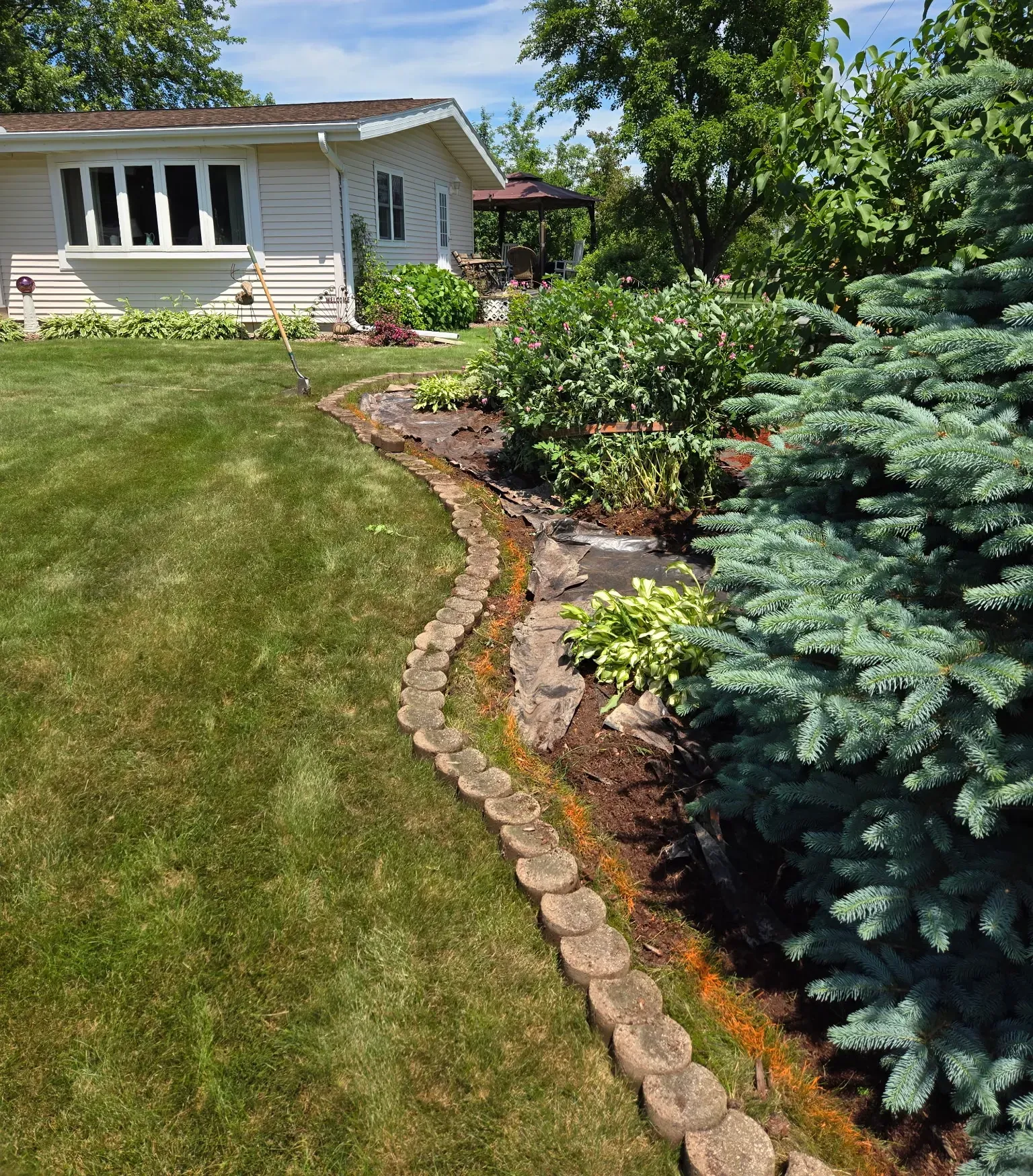 Concrete block edging being reset along a curved landscape bed with fabric and soil prep beside a blue spruce in Spencer, WI