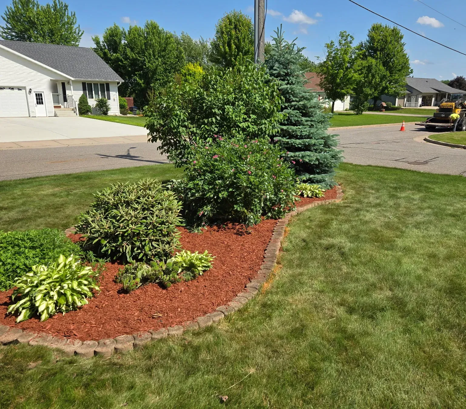 Curved landscape bed with fresh red mulch and reset block edging around blue spruce and flowering shrubs at a street corner in Spencer, WI