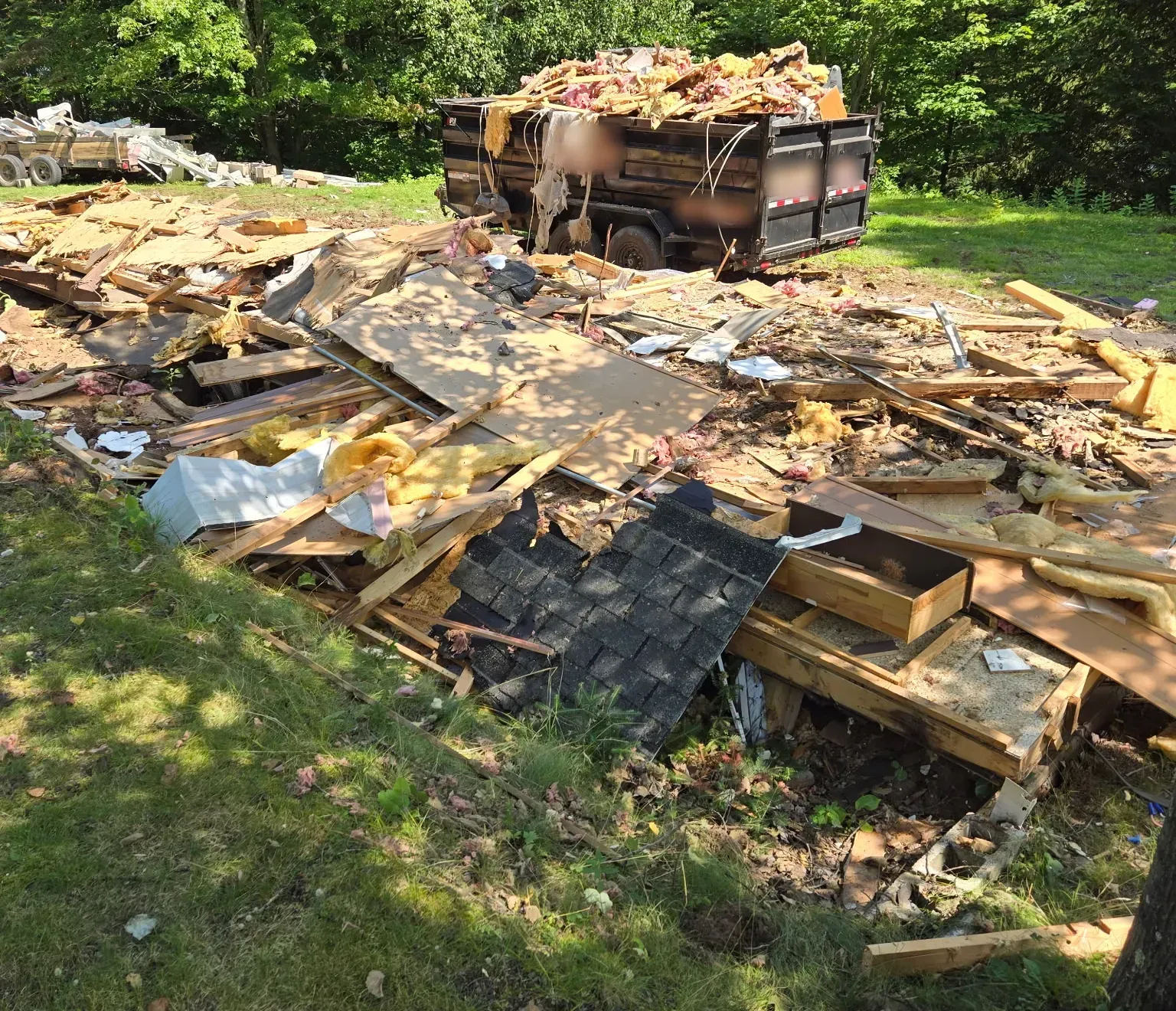 Field of demolished mobile home debris, including roof shingles, framing, insulation, and siding, beside a loaded dumpster in Phillips, WI