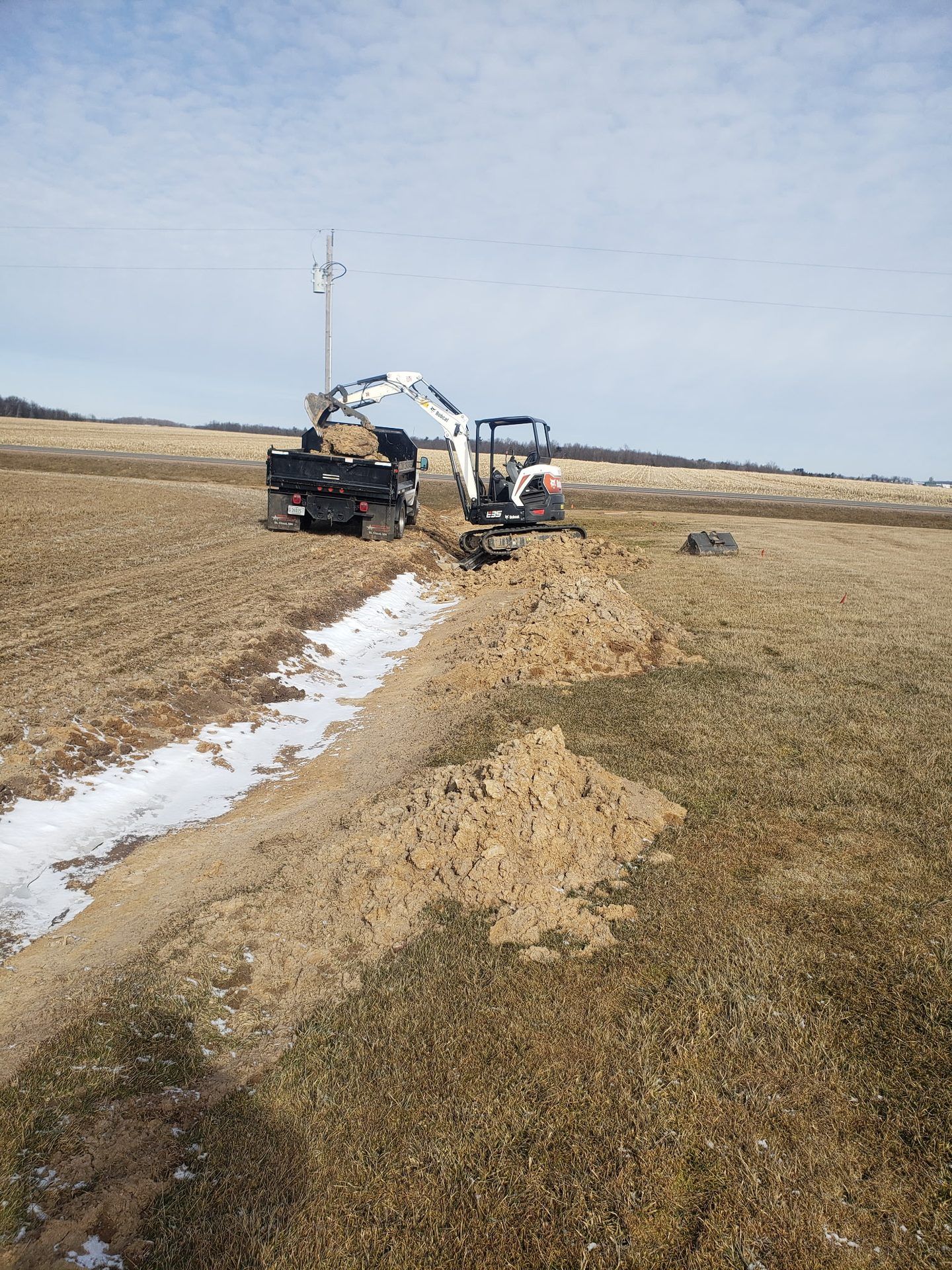 Excavator re-cutting a frozen field-edge ditch and hauling excavated soil by dump truck in Dorchester, WI