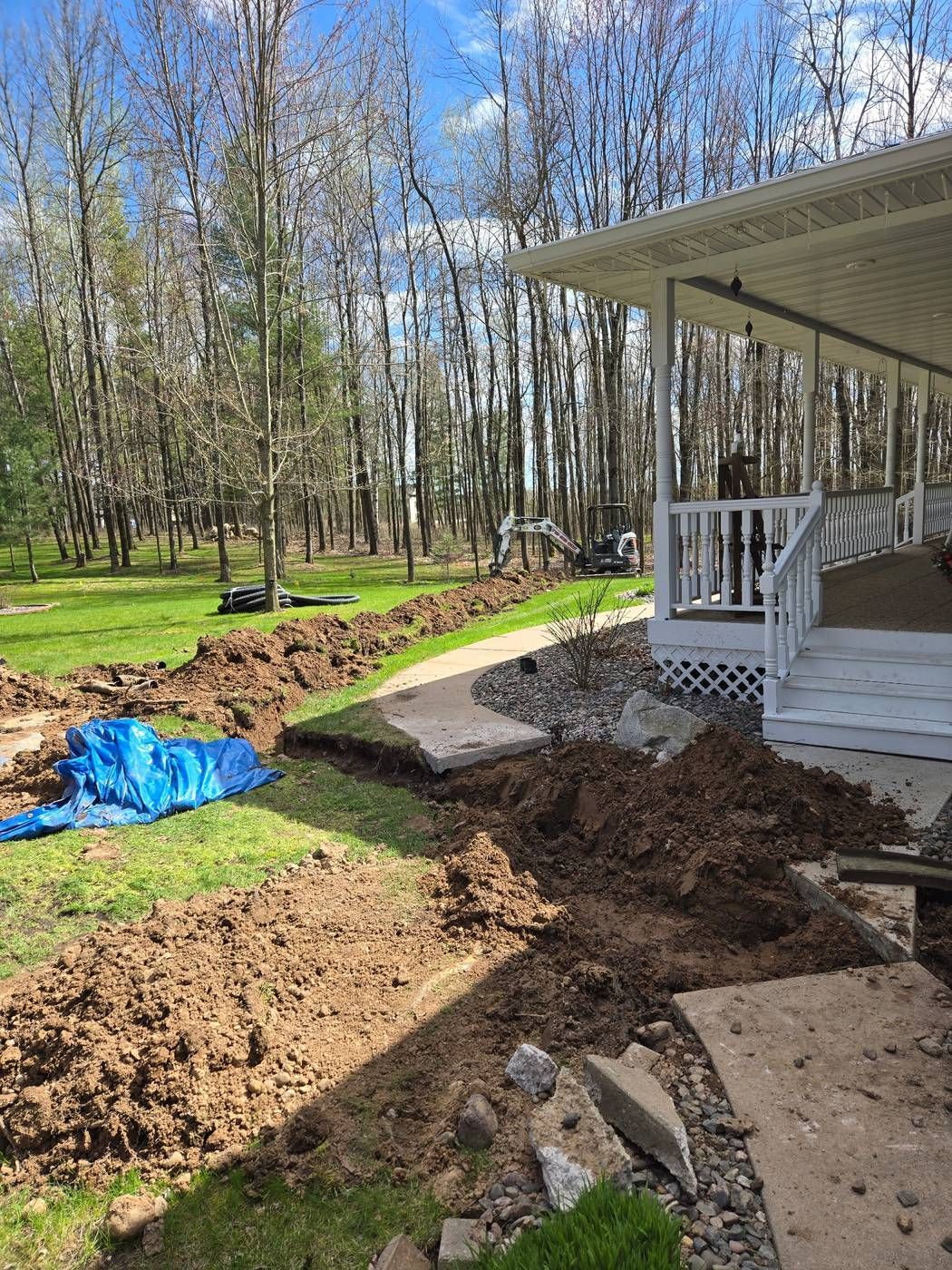 Drain tile trench running across a front yard and through a saw-cut walkway, with a mini excavator and rolls of drain pipe in the background in Medford, WI