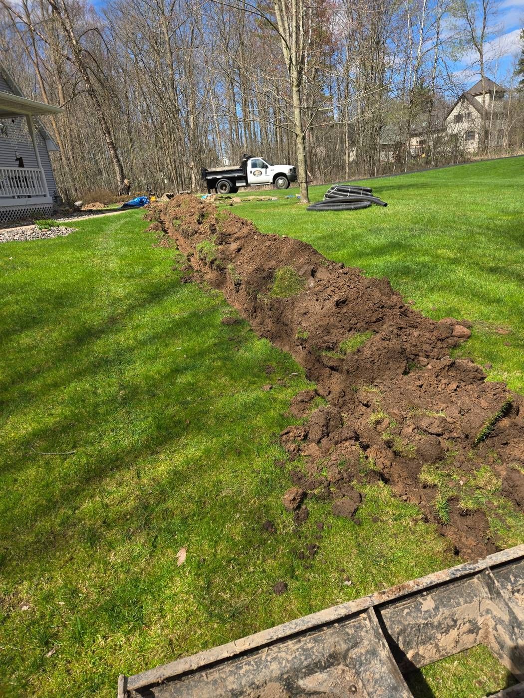 Long open trench dug across a residential lawn for drain tile installation, with the ERE dump truck and drain pipe coils nearby in Medford, WI