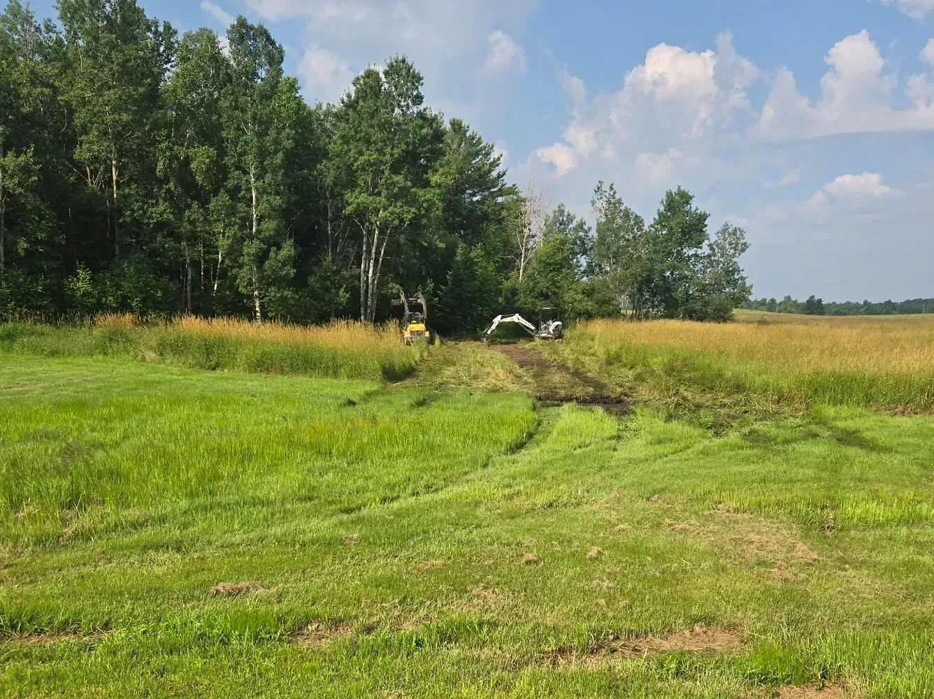 Freshly dug drainage ditch running from a mowed yard into tall grass toward a mini excavator and track loader at the tree line in Rib Lake, WI