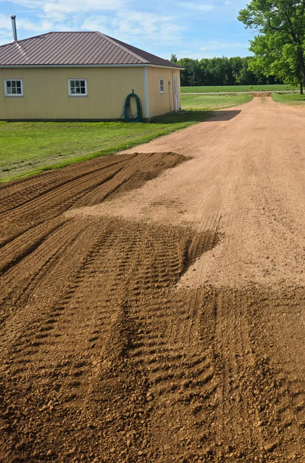 Fresh crushed red gravel top dressing spread over a worn farm driveway beside a yellow metal-roofed shop in Spencer, WI
