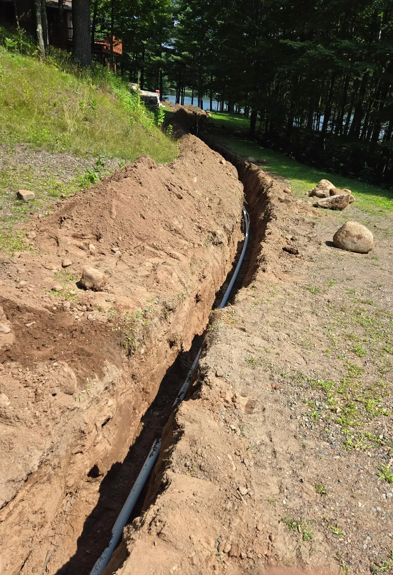 Deep trench with gray electrical conduit laid in the bottom, running downhill through a wooded lakefront lot in Phillips, WI