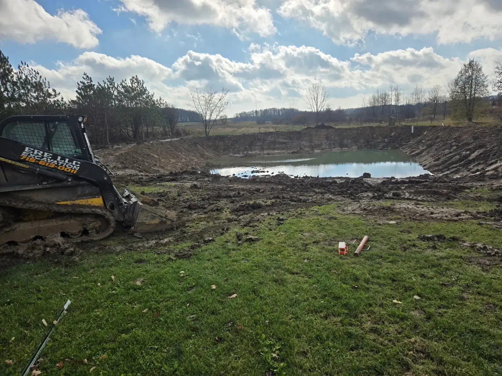 ERE LLC Excavation skid steer beside a freshly restored pond with reshaped, sloped banks refilling with water near Marshfield, WI
