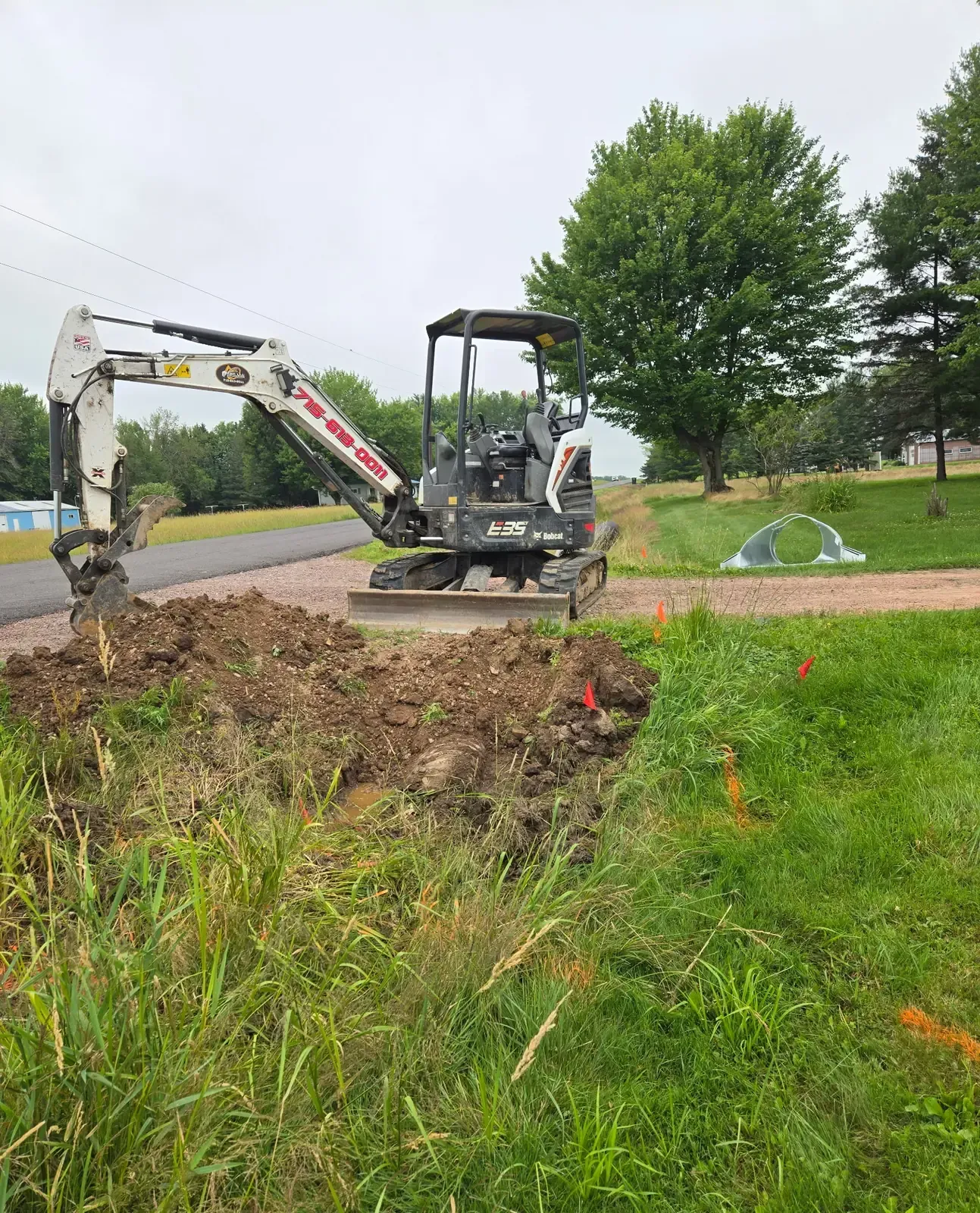 Mini excavator digging out an old driveway culvert with utility locate flags marked in Marshfield, WI.
