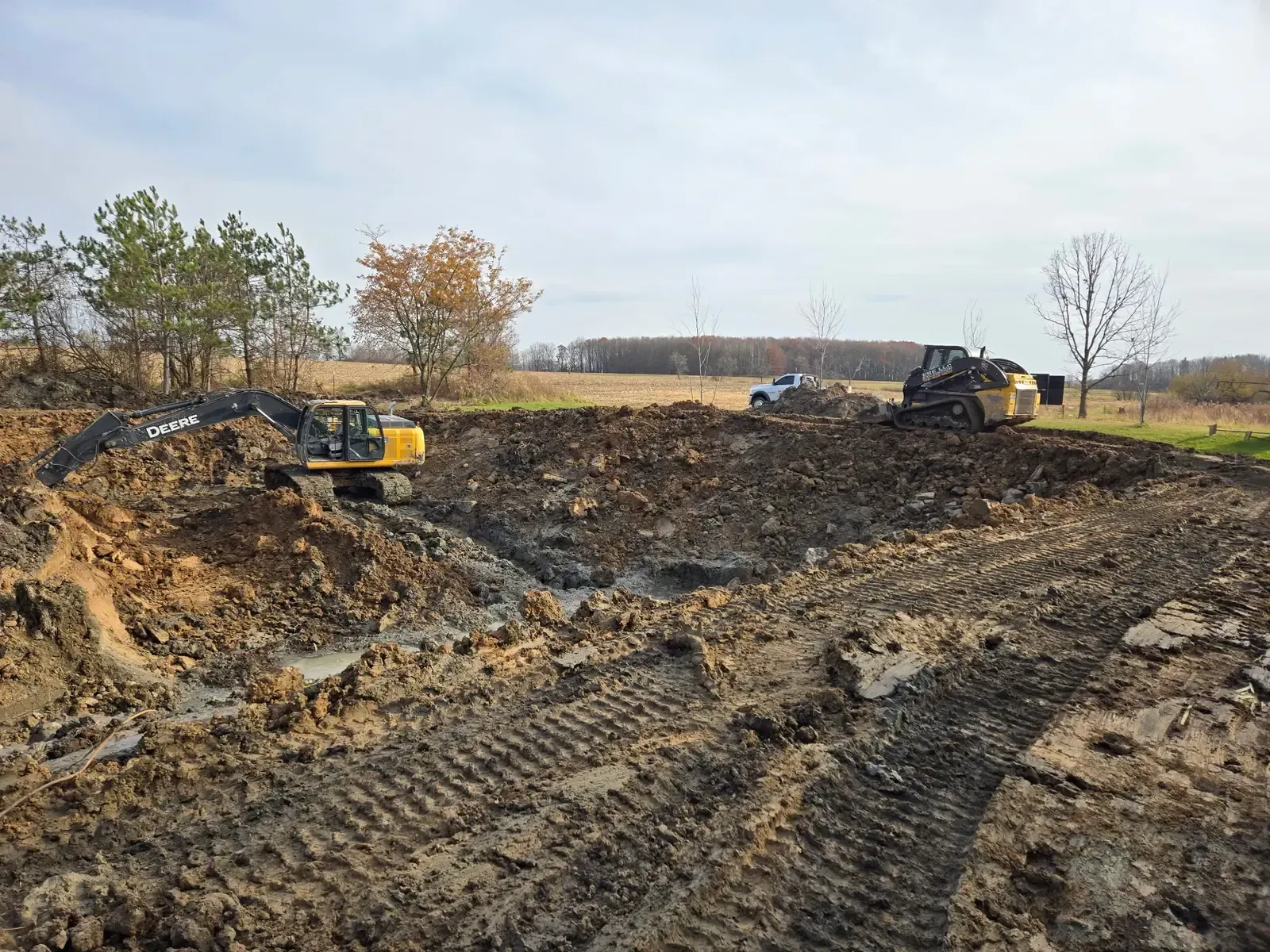 Excavator digging muck and sediment from a pond basin while an ERE LLC tracked skid steer moves spoils along the bank near Marshfield, WI