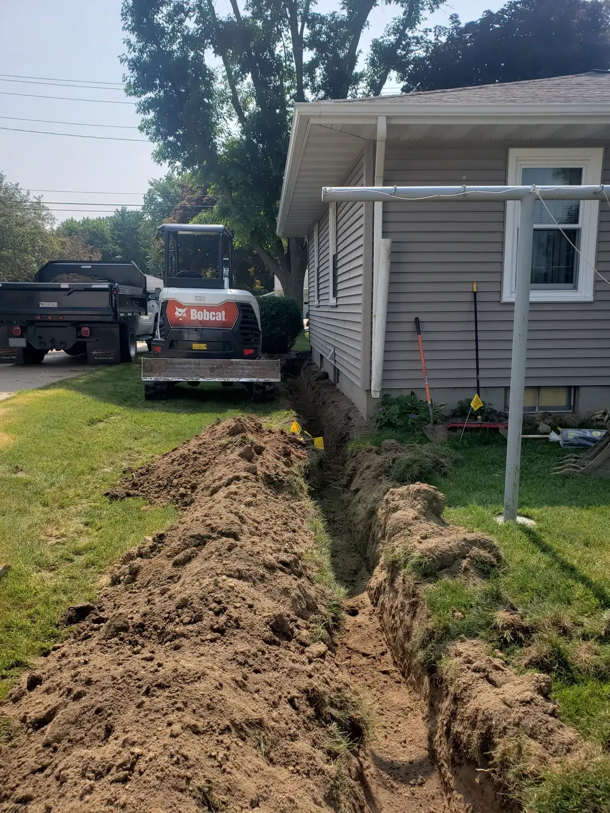 Discharge line trench running from a French drain toward the yard with mini excavator and dump truck in Marshfield, WI