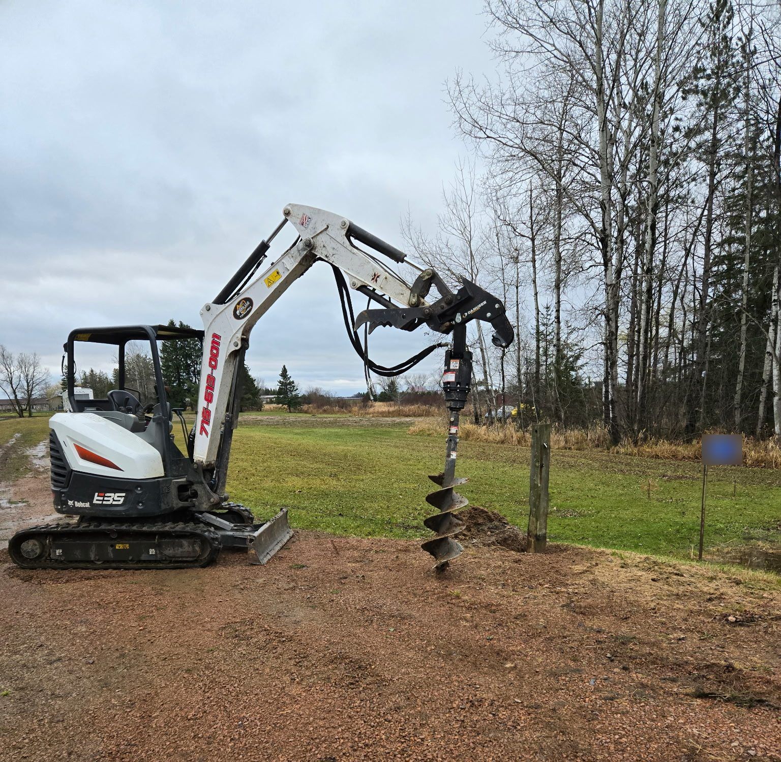 Bobcat E35 mini excavator with auger attachment beside a newly set wood post at a gravel driveway in Dorchester, WI